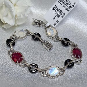 May include: A silver bracelet featuring oval-cut ruby and moonstone gemstones, interspersed with black onyx accents. The bracelet has a delicate design with a secure clasp. The gemstones are set in a silver setting with small, sparkling accents.