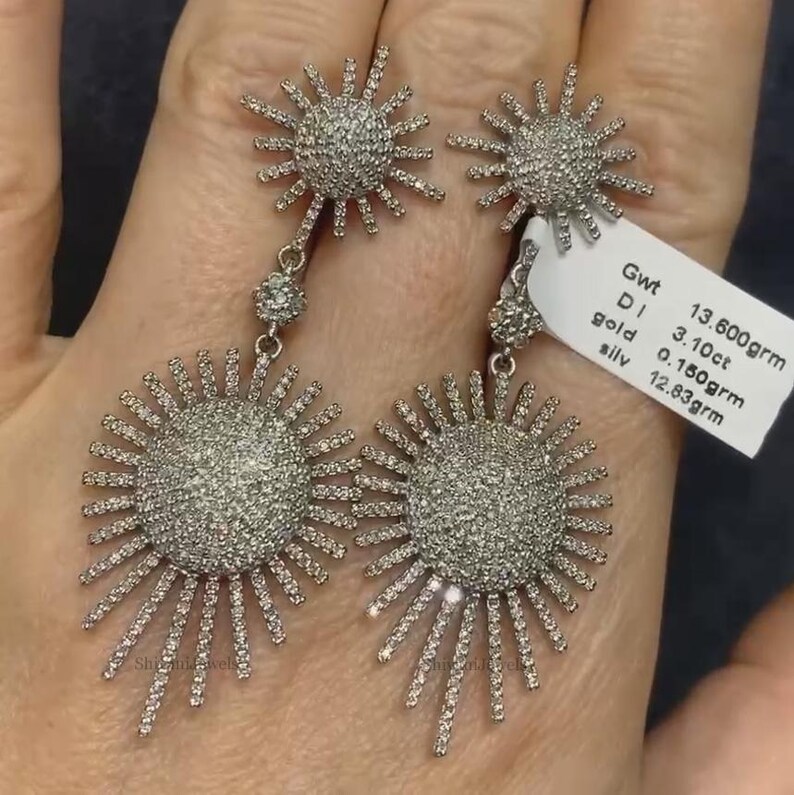 May include: A pair of silver earrings with a sunburst design. Each earring features a large, round, pave-set diamond center surrounded by smaller diamonds. The earrings are 13.600 grams in weight, with 3.10 carats of diamonds and 0.150 grams of gold. The earrings are also 12.83 grams of silver.