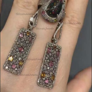 May include: A silver-toned jewelry set featuring a ring and matching earrings. The ring has a large, dark gemstone surrounded by smaller gems. The earrings are rectangular with colorful stones. The brand name "ShivaniJewels" is visible.
