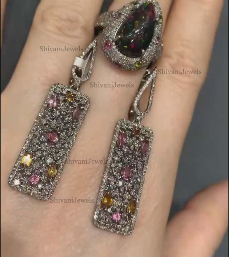 May include: A silver-toned jewelry set featuring a ring and matching earrings. The ring has a large, dark gemstone surrounded by smaller gems. The earrings are rectangular with colorful stones. The brand name "ShivaniJewels" is visible.