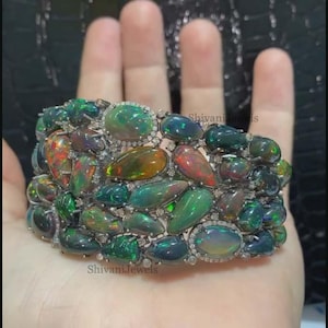 Puede incluir: Un brazalete negro y plateado con un gran grupo de ópalos verdes y negros. Los ópalos están engastados en una banda plateada y están rodeados de pequeños diamantes.