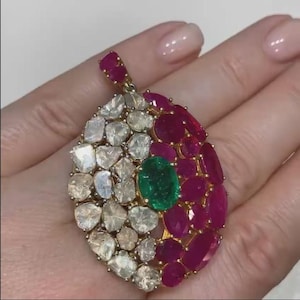 Puede incluir: Un colgante de oro con diseño de lágrima, con piedras preciosas blancas, rojo rubí y verde esmeralda. El colgante se sostiene en una mano, mostrando los intrincados detalles de la joyería.