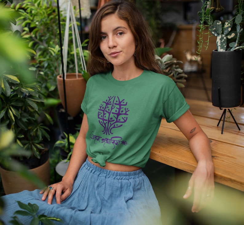 May include: A woman wearing a green t-shirt with a purple tree design and the text "সবুজ প্রকৃতির সঙ্গে" printed on it. She is sitting on a wooden surface with plants in the background.