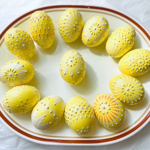 May include: A collection of 13 yellow Easter eggs with white and gold painted designs. The eggs are arranged in a circle on a white plate with a brown rim.