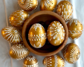 May include: A collection of golden Easter eggs decorated with white paint in various patterns. The eggs are arranged in a circle around a wooden bowl.