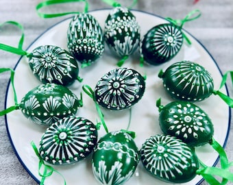 May include: A collection of green Easter eggs decorated with white intricate designs. The eggs are arranged on a white plate with a blue rim. Each egg has a unique pattern, including swirls, dots, and lines.