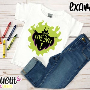Puede incluir: Camiseta blanca con un gr&aacute;fico verde y negro de un villano de dibujos animados con el texto "Long Live Evil!" impreso en ella. La camiseta est&aacute; colocada sobre una superficie de madera con un par de pantalones vaqueros azules y l&aacute;pices de colores.