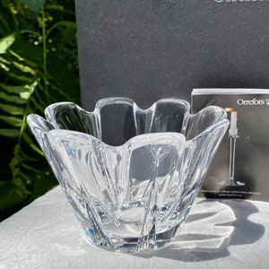 Puede incluir: Un cuenco de vidrio transparente con un borde festoneado. El cuenco está hecho por Orrefors, un fabricante de vidrio sueco.