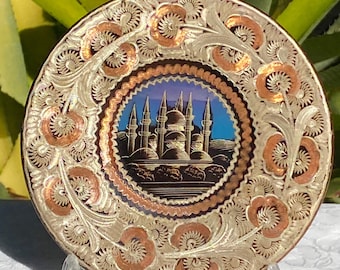 May include: A small, round, copper and silver plate with an intricate floral design. The center of the plate features a painted image of a mosque with minarets against a blue sky.