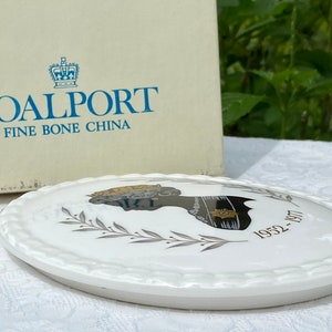 A vintage oval wall plaque that has a silhouette of Queen Elizabeth II.  The cameo style silhouette is black with gold accents and has the dates 1952-1977 underneath her figure.  The back of the plaque reads "Coalport Made in England - Her Majesty Queen Elizabeth II Silver Jubilee 1952-1977 - Silhouette by LJ Arnold."  It has two holes at the top to run a wire for hanging.  Has its original box with original packaging.  Circa 1970s. No damage. Box shows signs of shelf wear. Measures 6" x 4 3/4"