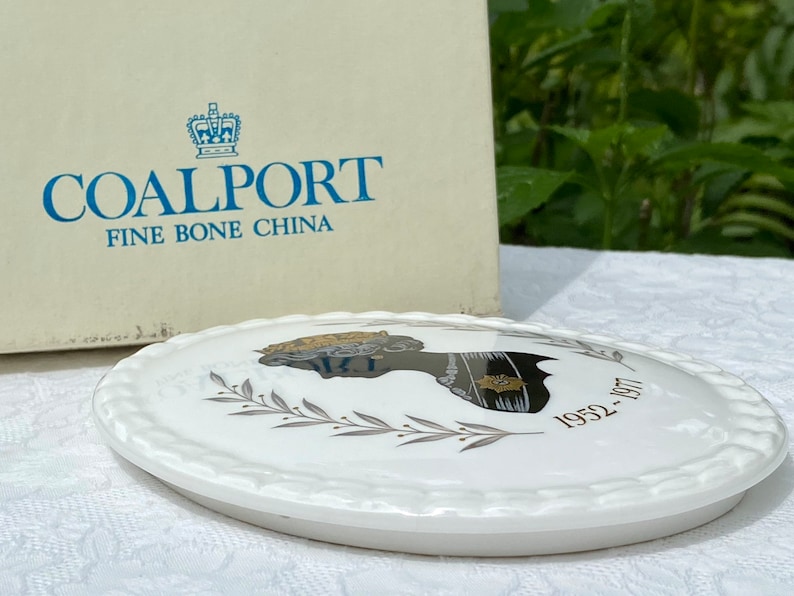 A vintage oval wall plaque that has a silhouette of Queen Elizabeth II.  The cameo style silhouette is black with gold accents and has the dates 1952-1977 underneath her figure.  The back of the plaque reads "Coalport Made in England - Her Majesty Queen Elizabeth II Silver Jubilee 1952-1977 - Silhouette by LJ Arnold."  It has two holes at the top to run a wire for hanging.  Has its original box with original packaging.  Circa 1970s. No damage. Box shows signs of shelf wear. Measures 6" x 4 3/4"