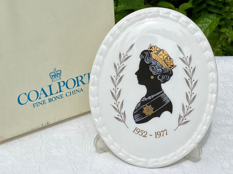 A vintage oval wall plaque that has a silhouette of Queen Elizabeth II.  The cameo style silhouette is black with gold accents and has the dates 1952-1977 underneath her figure.  The back of the plaque reads "Coalport Made in England - Her Majesty Queen Elizabeth II Silver Jubilee 1952-1977 - Silhouette by LJ Arnold."  It has two holes at the top to run a wire for hanging.  Has its original box with original packaging.  Circa 1970s. No damage. Box shows signs of shelf wear. Measures 6" x 4 3/4"