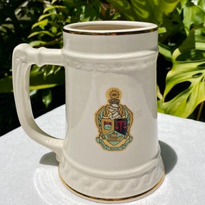 May include: White ceramic beer stein with a gold rim and a crest featuring a shield with a scale of justice, a book, and a ship. The crest is surrounded by a wreath and a banner that reads "Kappa Sigma".