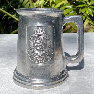 May include: A silver pewter beer stein with a raised embossed design of a lion and a crown with the text "STROH'S" underneath.