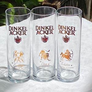 May include: Three clear glass beer glasses with the text "Dinkel Acker" printed on the front. Each glass features a different zodiac symbol: Gemini, Leo, and Sagittarius.