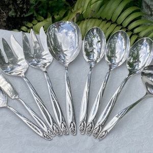May include: A set of nine silver serving utensils with ornate handles. The utensils include a butter knife, a sugar spoon, two serving forks, a slotted spoon, a ladle, and two serving spoons.