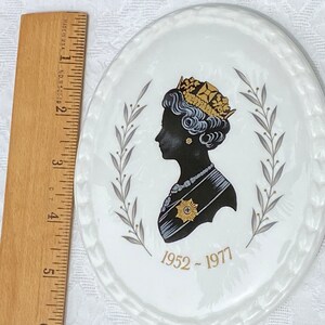 A vintage oval wall plaque that has a silhouette of Queen Elizabeth II.  The cameo style silhouette is black with gold accents and has the dates 1952-1977 underneath her figure.  It is next to a wooden ruler. It measures 6 inches x 4 3/4 inches.