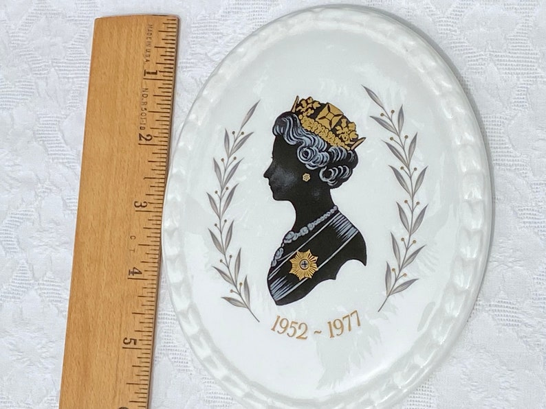 A vintage oval wall plaque that has a silhouette of Queen Elizabeth II.  The cameo style silhouette is black with gold accents and has the dates 1952-1977 underneath her figure.  It is next to a wooden ruler. It measures 6 inches x 4 3/4 inches.