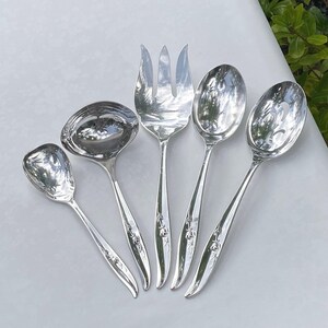 May include: A set of five silver serving utensils, including a ladle, serving fork, and spoons. The utensils have a reflective surface and floral detailing on the handles. The arrangement is on a white surface.