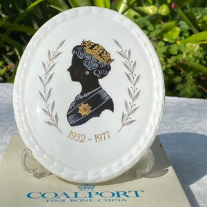 A vintage oval wall plaque that has a silhouette of Queen Elizabeth II.  The cameo style silhouette is black with gold accents and has the dates 1952-1977 underneath her figure.  The back of the plaque reads "Coalport Made in England - Her Majesty Queen Elizabeth II Silver Jubilee 1952-1977 - Silhouette by LJ Arnold."  It has two holes at the top to run a wire for hanging.  Has its original box with original packaging.  Circa 1970s. No damage. Box shows signs of shelf wear. Measures 6" x 4 3/4"