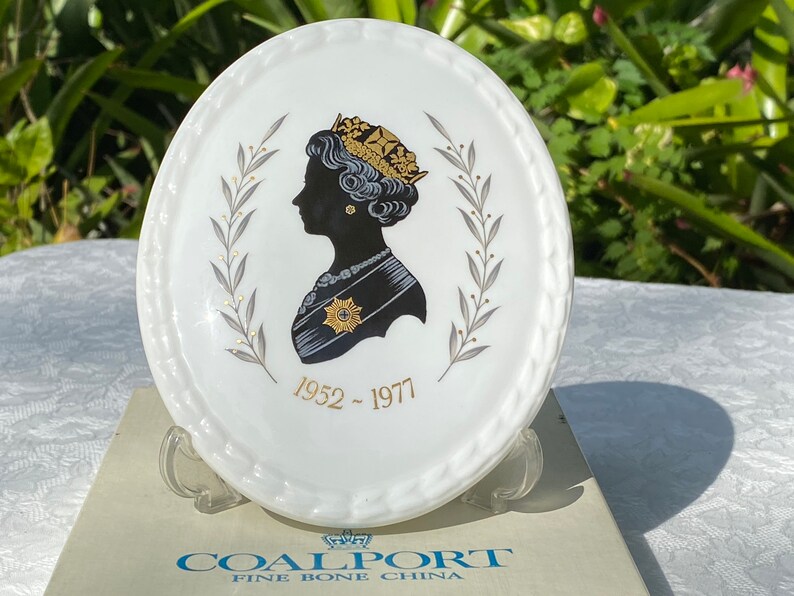 A vintage oval wall plaque that has a silhouette of Queen Elizabeth II.  The cameo style silhouette is black with gold accents and has the dates 1952-1977 underneath her figure.  The back of the plaque reads "Coalport Made in England - Her Majesty Queen Elizabeth II Silver Jubilee 1952-1977 - Silhouette by LJ Arnold."  It has two holes at the top to run a wire for hanging.  Has its original box with original packaging.  Circa 1970s. No damage. Box shows signs of shelf wear. Measures 6" x 4 3/4"