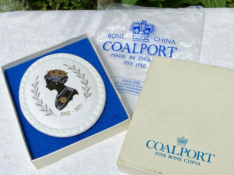 A vintage oval wall plaque that has a silhouette of Queen Elizabeth II.  The cameo style silhouette is black with gold accents and has the dates 1952-1977 underneath her figure.  The back of the plaque reads "Coalport Made in England - Her Majesty Queen Elizabeth II Silver Jubilee 1952-1977 - Silhouette by LJ Arnold."  It has two holes at the top to run a wire for hanging.  Has its original box with original packaging.  Circa 1970s. No damage. Box shows signs of shelf wear. Measures 6" x 4 3/4"