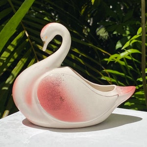 May include: A white ceramic swan planter with a pink ombre glaze. The swan has a long neck and a small opening for planting.