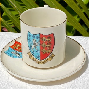May include: A white porcelain teacup and saucer with a gold trim. The cup features a red and blue coat of arms with a lion and the word "Shoreham".