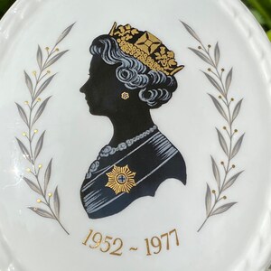 A vintage oval wall plaque that has a silhouette of Queen Elizabeth II.  The cameo style silhouette is black with gold accents and has the dates 1952-1977 underneath her figure.  The back of the plaque reads "Coalport Made in England - Her Majesty Queen Elizabeth II Silver Jubilee 1952-1977 - Silhouette by LJ Arnold."  It has two holes at the top to run a wire for hanging.  Has its original box with original packaging.  Circa 1970s. No damage. Box shows signs of shelf wear. Measures 6" x 4 3/4"