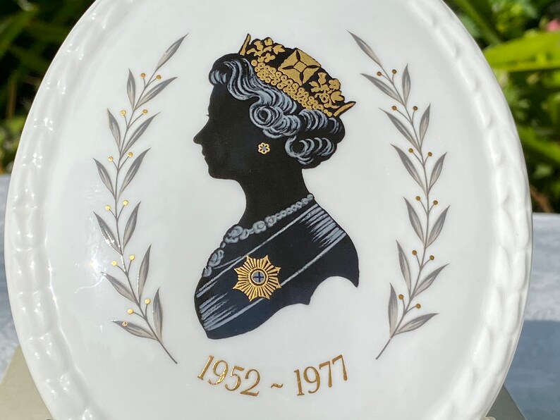 A vintage oval wall plaque that has a silhouette of Queen Elizabeth II.  The cameo style silhouette is black with gold accents and has the dates 1952-1977 underneath her figure.  The back of the plaque reads "Coalport Made in England - Her Majesty Queen Elizabeth II Silver Jubilee 1952-1977 - Silhouette by LJ Arnold."  It has two holes at the top to run a wire for hanging.  Has its original box with original packaging.  Circa 1970s. No damage. Box shows signs of shelf wear. Measures 6" x 4 3/4"