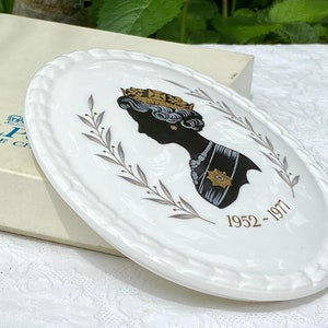 A vintage oval wall plaque that has a silhouette of Queen Elizabeth II.  The cameo style silhouette is black with gold accents and has the dates 1952-1977 underneath her figure.  The back of the plaque reads "Coalport Made in England - Her Majesty Queen Elizabeth II Silver Jubilee 1952-1977 - Silhouette by LJ Arnold."  It has two holes at the top to run a wire for hanging.  Has its original box with original packaging.  Circa 1970s. No damage. Box shows signs of shelf wear. Measures 6" x 4 3/4"