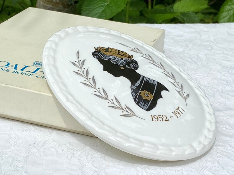 A vintage oval wall plaque that has a silhouette of Queen Elizabeth II.  The cameo style silhouette is black with gold accents and has the dates 1952-1977 underneath her figure.  The back of the plaque reads "Coalport Made in England - Her Majesty Queen Elizabeth II Silver Jubilee 1952-1977 - Silhouette by LJ Arnold."  It has two holes at the top to run a wire for hanging.  Has its original box with original packaging.  Circa 1970s. No damage. Box shows signs of shelf wear. Measures 6" x 4 3/4"