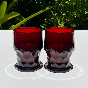 May include: Two ruby red glass tumblers with a textured, faceted design. The glasses are empty and sitting on a white surface.