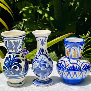May include: Three small ceramic vases with blue and white painted designs. The vase on the left has a handle and a floral pattern. The middle vase has a windmill design. The vase on the right has a geometric pattern with blue, white, and gold accents.