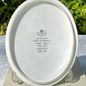 A vintage oval wall plaque that has a silhouette of Queen Elizabeth II.  The cameo style silhouette is black with gold accents and has the dates 1952-1977 underneath her figure.  The back of the plaque reads "Coalport Made in England - Her Majesty Queen Elizabeth II Silver Jubilee 1952-1977 - Silhouette by LJ Arnold."  It has two holes at the top to run a wire for hanging.  Has its original box with original packaging.  Circa 1970s. No damage. Box shows signs of shelf wear. Measures 6" x 4 3/4"