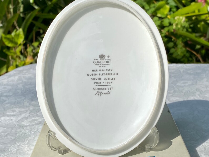 A vintage oval wall plaque that has a silhouette of Queen Elizabeth II.  The cameo style silhouette is black with gold accents and has the dates 1952-1977 underneath her figure.  The back of the plaque reads "Coalport Made in England - Her Majesty Queen Elizabeth II Silver Jubilee 1952-1977 - Silhouette by LJ Arnold."  It has two holes at the top to run a wire for hanging.  Has its original box with original packaging.  Circa 1970s. No damage. Box shows signs of shelf wear. Measures 6" x 4 3/4"