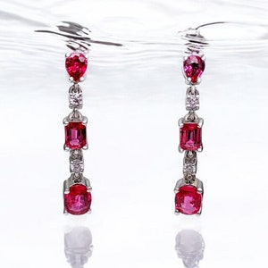 以下が含まれることがあります： 赤い宝石と小さな透明なダイヤモンドが付いた、ぶら下がっているイヤリングのペア。イヤリングは銀のポストから吊り下げられています。