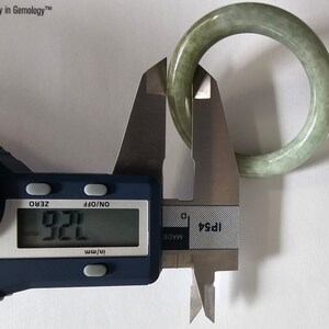 以下が含まれることがあります： デジタルノギスで測定されている翡翠のバングル。ノギスは7.21mmを示しています。バングルは薄い緑色です。