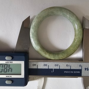 以下が含まれることがあります： デジタルノギスで測定されている翡翠のバングルブレスレット。ノギスは48.02mmを示しています。バングルは薄い緑色で、滑らかで磨かれた表面をしています。