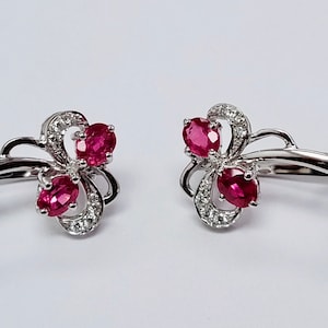 Op de afbeelding: Een paar zilveren oorbellen met een bloemmotief met drie rode robijnstenen en kleine witte diamanten.