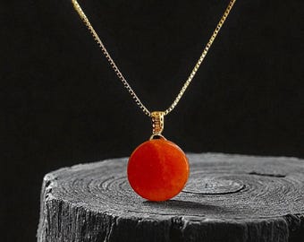 May include: A gold chain necklace featuring a circular, orange pendant. The pendant is a solid, round shape. The necklace is presented against a dark, textured backdrop, highlighting the pendant's vibrant colour.