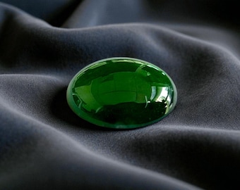 May include: A vibrant, oval-shaped, emerald-green gemstone rests on a dark gray fabric. The polished surface of the gem reflects light, creating highlights and shadows. The stone's smooth, rounded form contrasts with the textured background.
