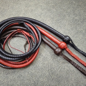 Puede incluir: Tres látigos de cuero, uno rojo y dos negros, están trenzados y enrollados juntos. Los látigos tienen un mango anudado.