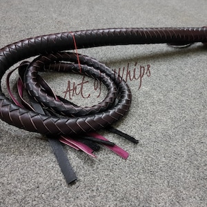 May include: A black leather bullwhip with a braided design and a pink and black tassel. The whip is coiled on a gray surface. The text "Art of Whips" is visible in the image.