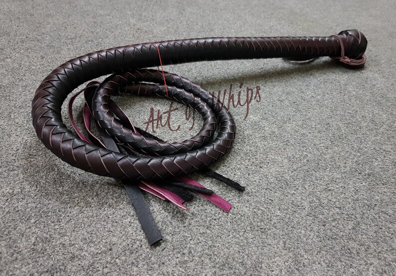 May include: A black leather bullwhip with a braided design and a pink and black tassel. The whip is coiled on a gray surface. The text "Art of Whips" is visible in the image.