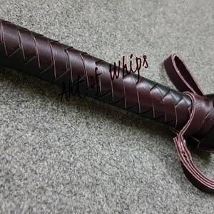 May include: A close-up of a dark brown leather whip with a braided handle and a loop at the end. The whip is made by Art of Whips.