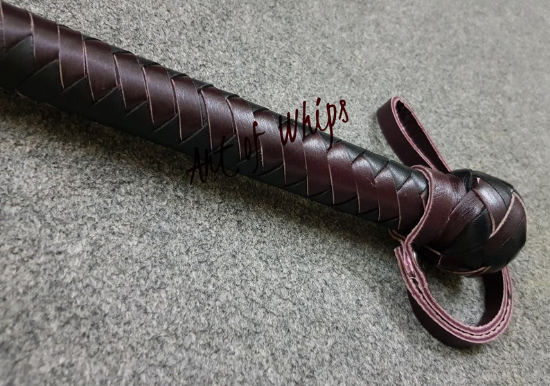May include: A close-up of a dark brown leather whip with a braided handle and a loop at the end. The whip is made by Art of Whips.