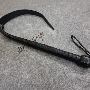 Puede incluir: Un l&aacute;tigo de cuero negro con un mango trenzado y una correa larga y fina. El l&aacute;tigo est&aacute; sobre una superficie gris. Art of Whips.