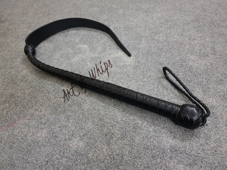 Puede incluir: Un l&aacute;tigo de cuero negro con un mango trenzado y una correa larga y fina. El l&aacute;tigo est&aacute; sobre una superficie gris. Art of Whips.