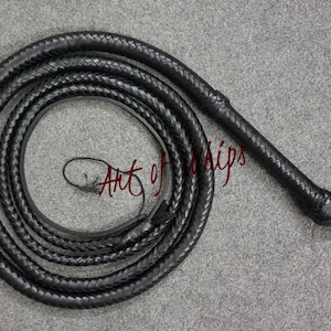 Puede incluir: Un látigo de cuero negro con un mango trenzado y un lazo en el extremo. El látigo está enrollado y colocado sobre una superficie gris. El texto "Art of Whips" es visible en la imagen.